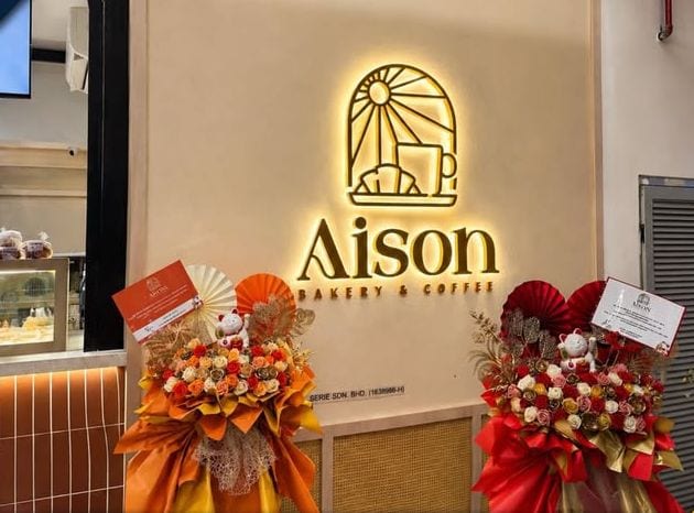 A backlit Aison Bakery and Coffee logo sign above a bakery counter on opening day, flanked by congratulation flower stands.