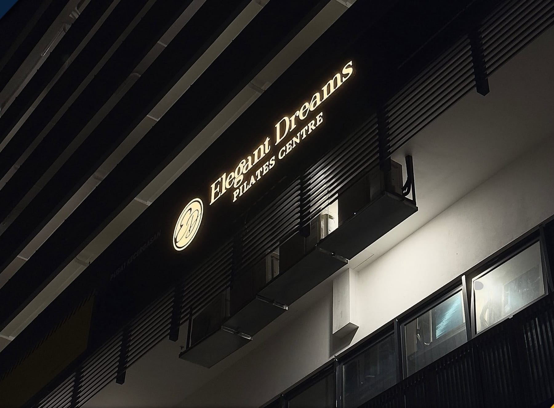Illuminated "Elegant Dreams Pilates Centre" lettering mounted high on a dark slatted building facade at dusk.