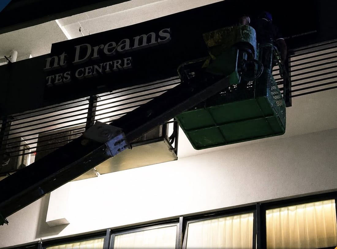 Night installation in progress: part of a sign already mounted on a dark slatted facade, with a worker at height beside it under work lighting.